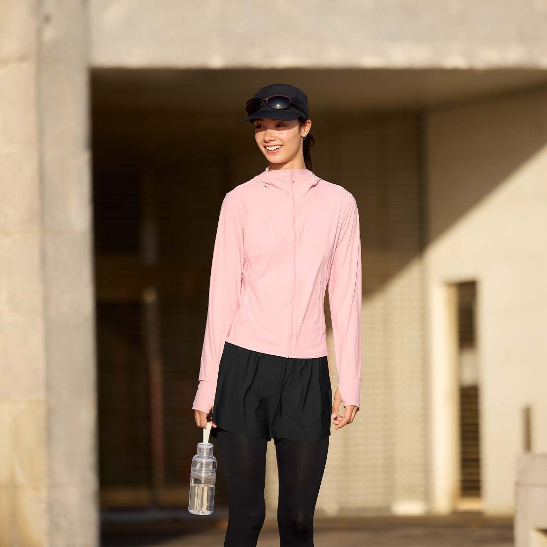 model wearing Ultra Stretch AIRism UV Protection Full-Zip Hoodie in pink with black shorts worn over black running tights, a black cap and sunglasses, while carrying a water bottle