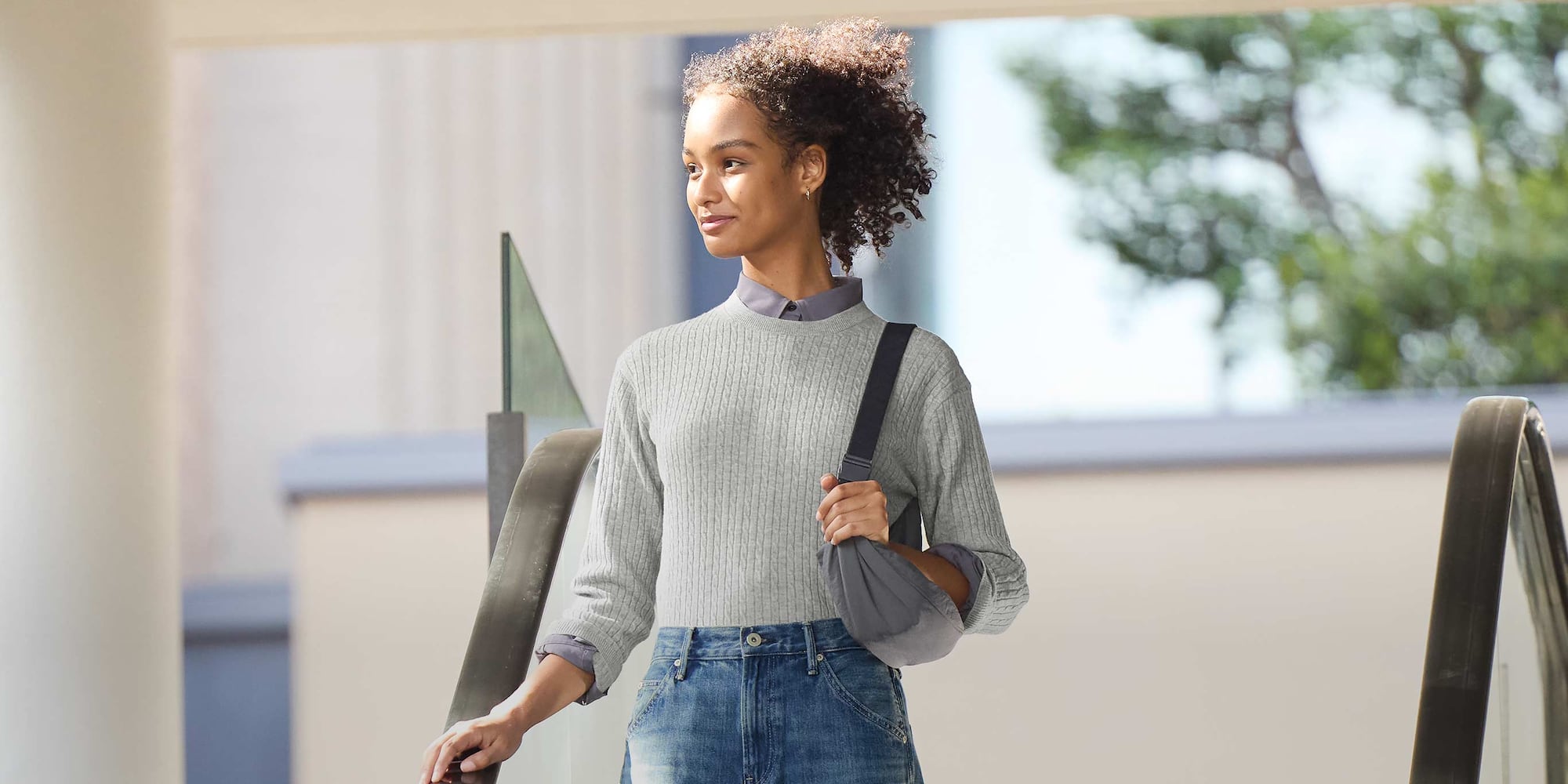 model wearing Mini Cable Crew Neck Jumper in grey over a dark grey blouse and jeans while carrying a dark grey round mini shoulder bag