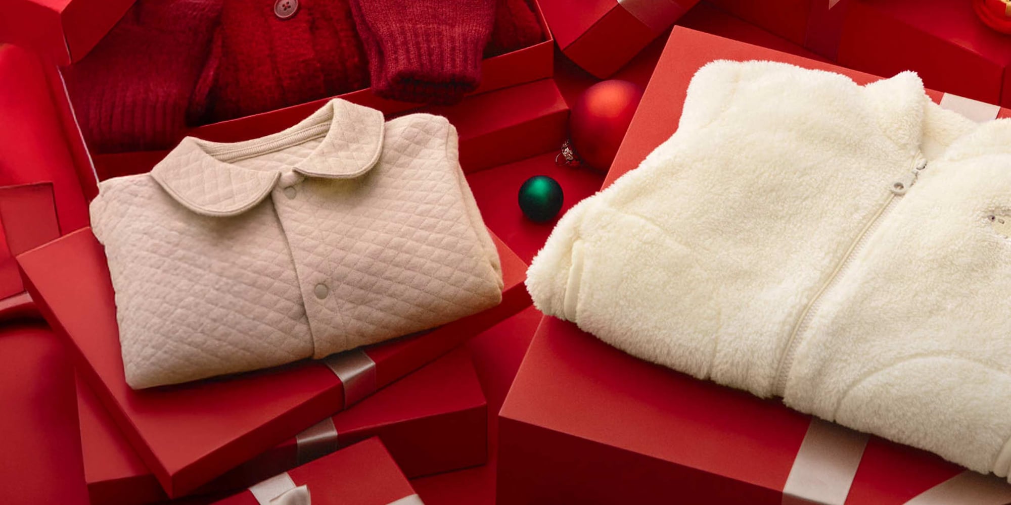 an arrangement including a knitted and quilted cardigan and an insulated one piece outfit against a red background with gift boxes, baubles and ribbon