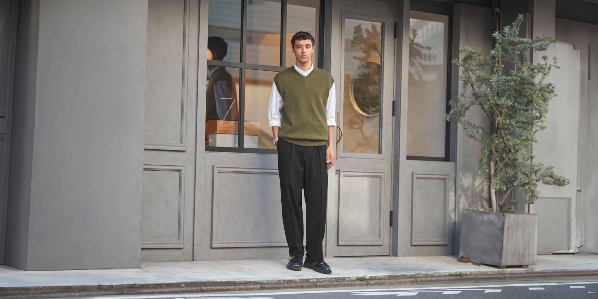 model wearing olive sweater vest over a white shirt with black wide leg trousers and black shoes