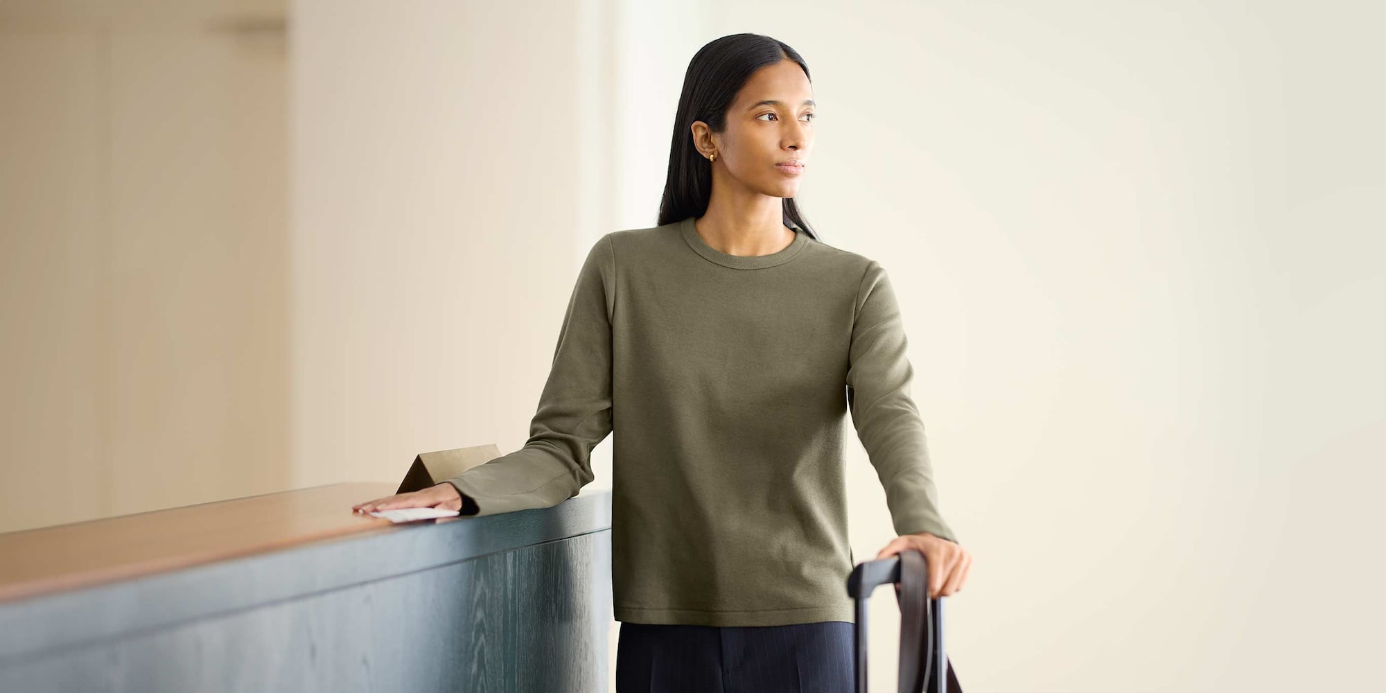 model at a hotel wearing a muted green smooth cotton long sleeve T-shirt with her hand on a suitcase