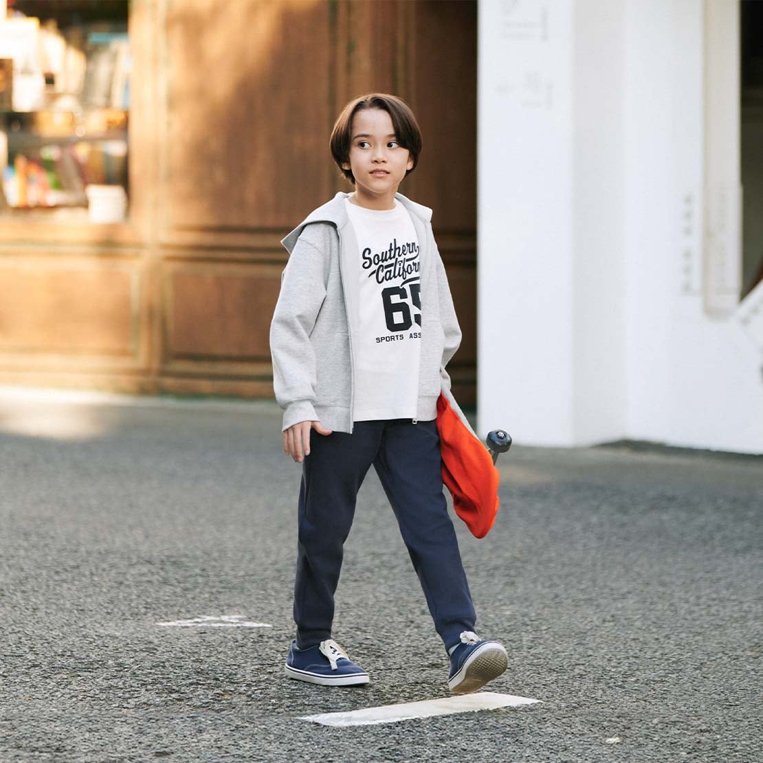 model wearing DRY Sweat Joggers in navy with a white graphic T-shirt and grey hoodie and blue trainers while carrying a skateboard