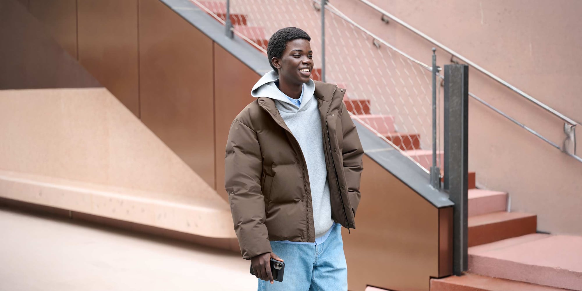 model wearing Seamless Down Jacket in brown over a light blue shirt, grey hoodie and light wash jeans, holding a smartphone