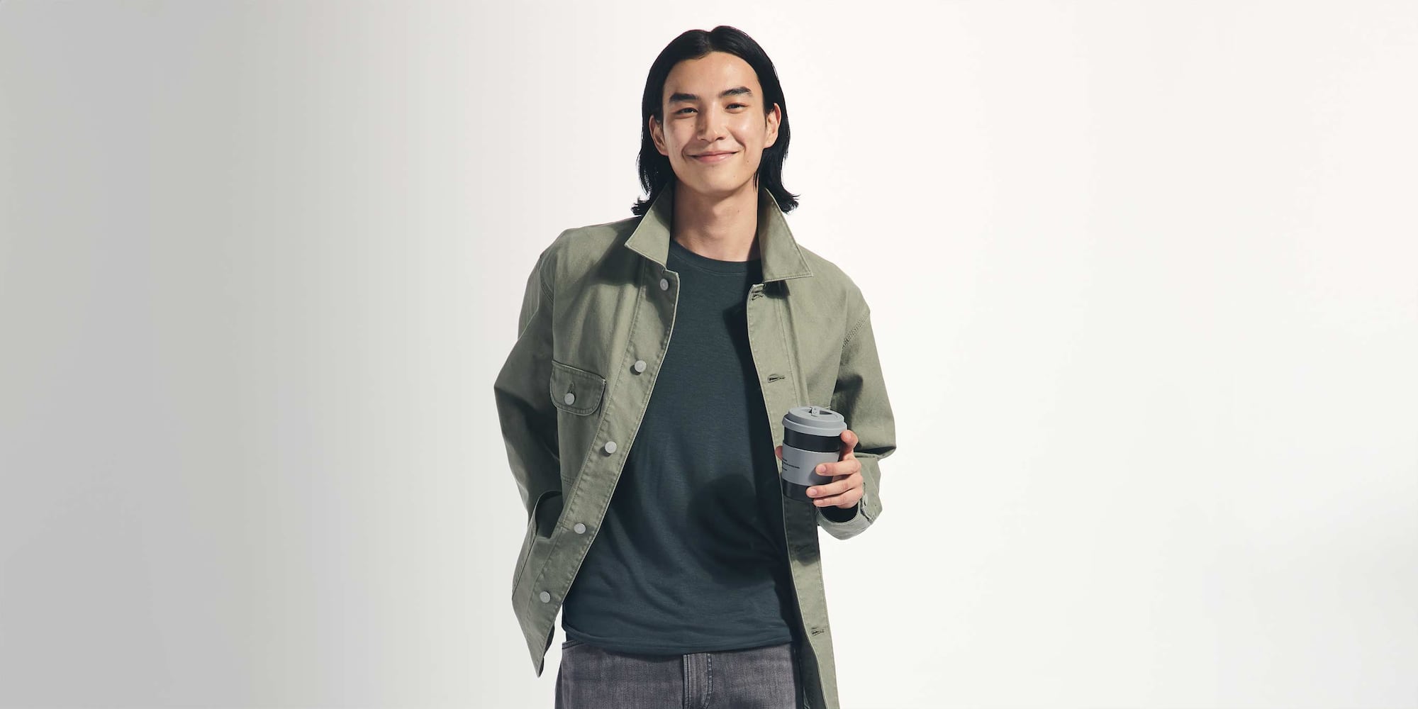 model wearing HEATTECH Extra Warm Cashmere Blend Crew Neck T-Shirt in dark green under a light green jacket with grey jeans while holding a reusable to-go coffee cup