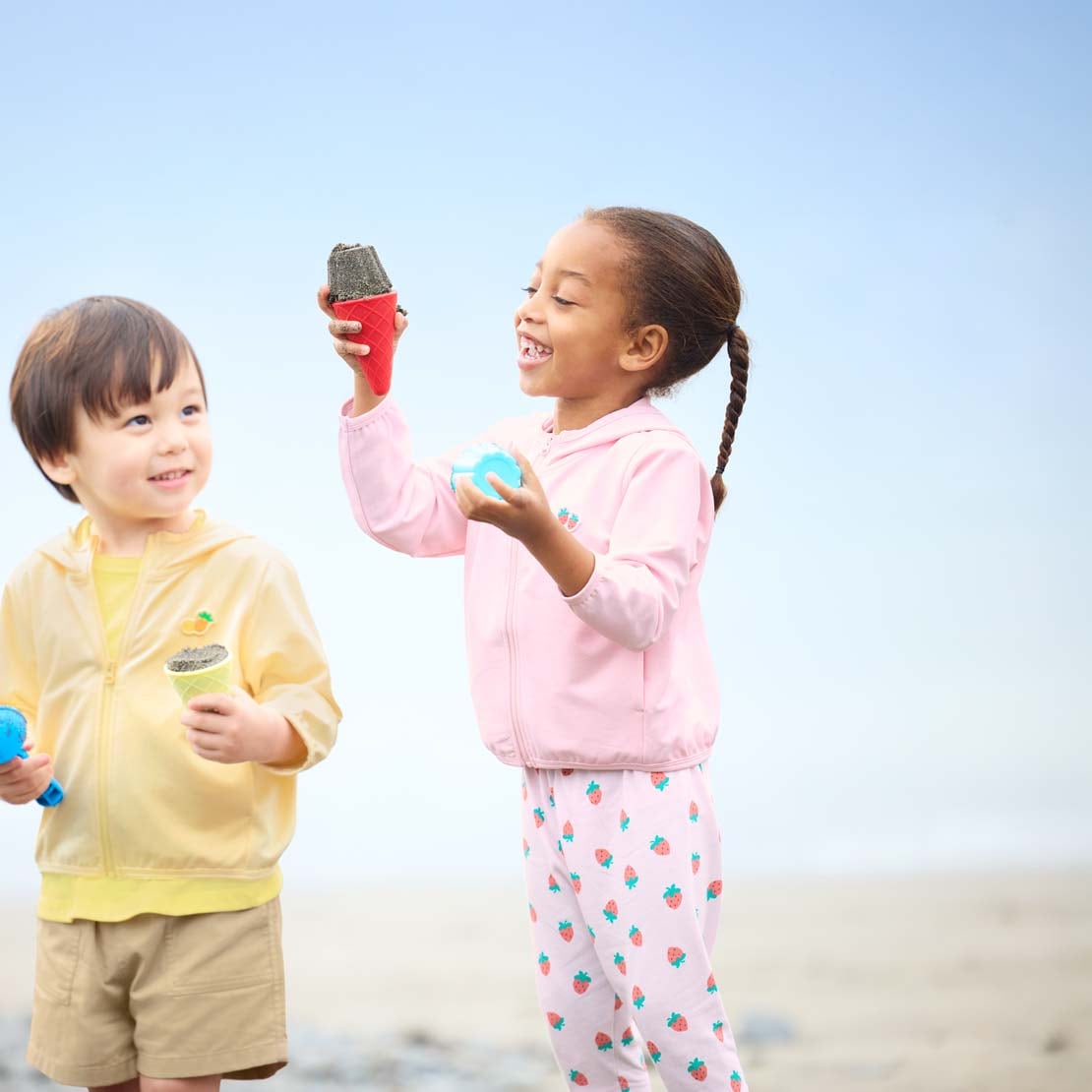 Due bimbi indossano felpe protezione UV con cappuccio nei colori giallo e rosa mentre giocano con la sabbia in spiaggia