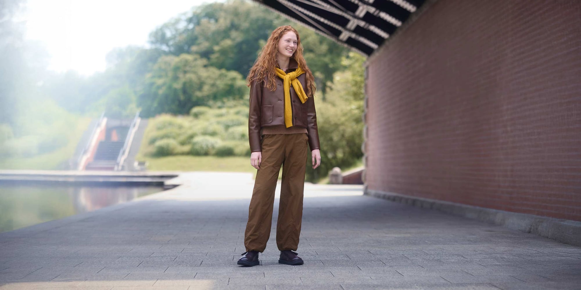 model wearing Warm Lined Trousers in brown with a brown jacket and shoes and a yellow knit tied around the shoulders, while standing underneath a bridge