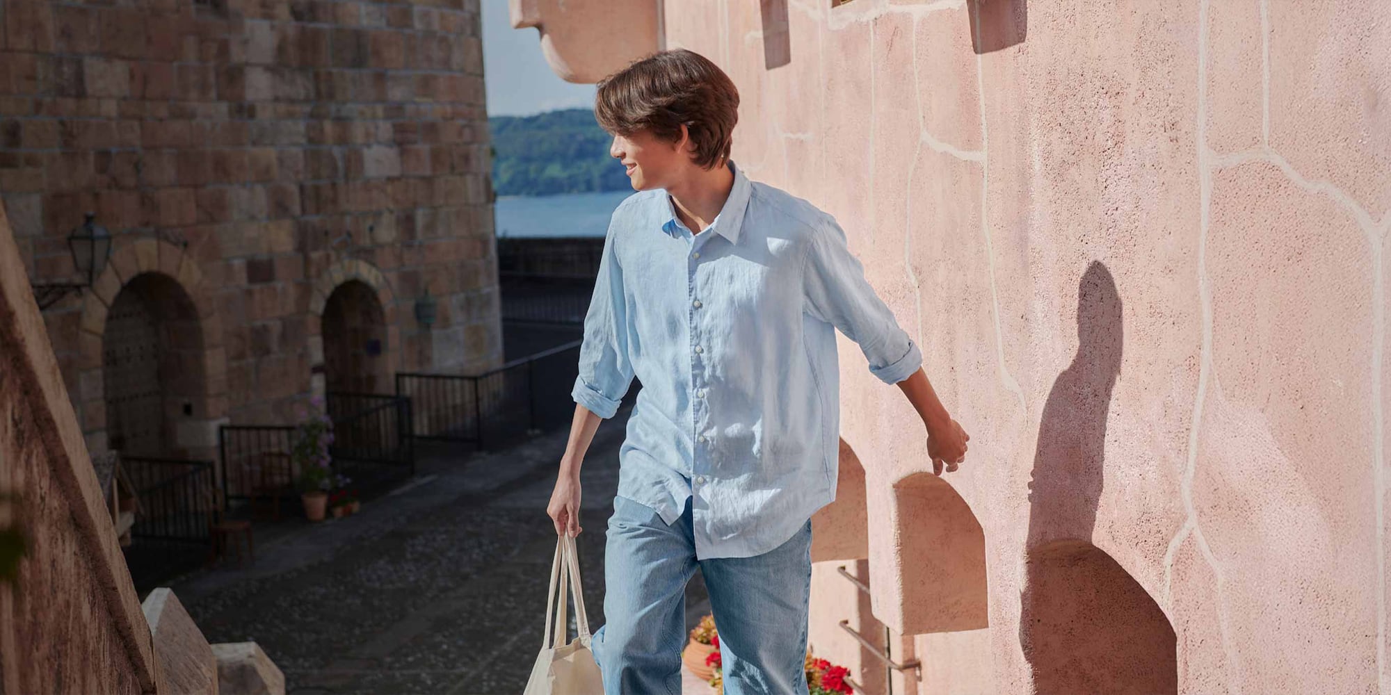 model wearing 100% Premium Linen Shirt in blue with mid wash jeans, carrying a tote bag with the sea in the background
