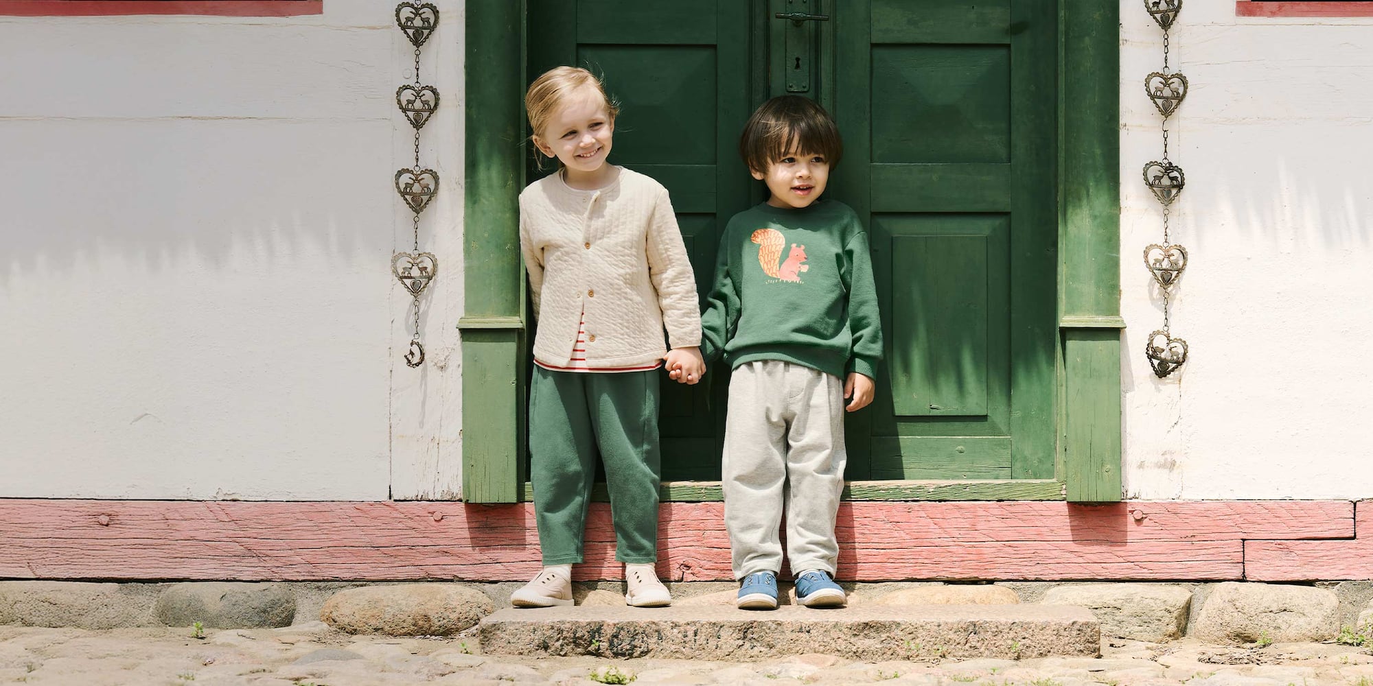 two models holding hands wearing a cream jacket and green trousers and a green sweatshirt and light grey trousers