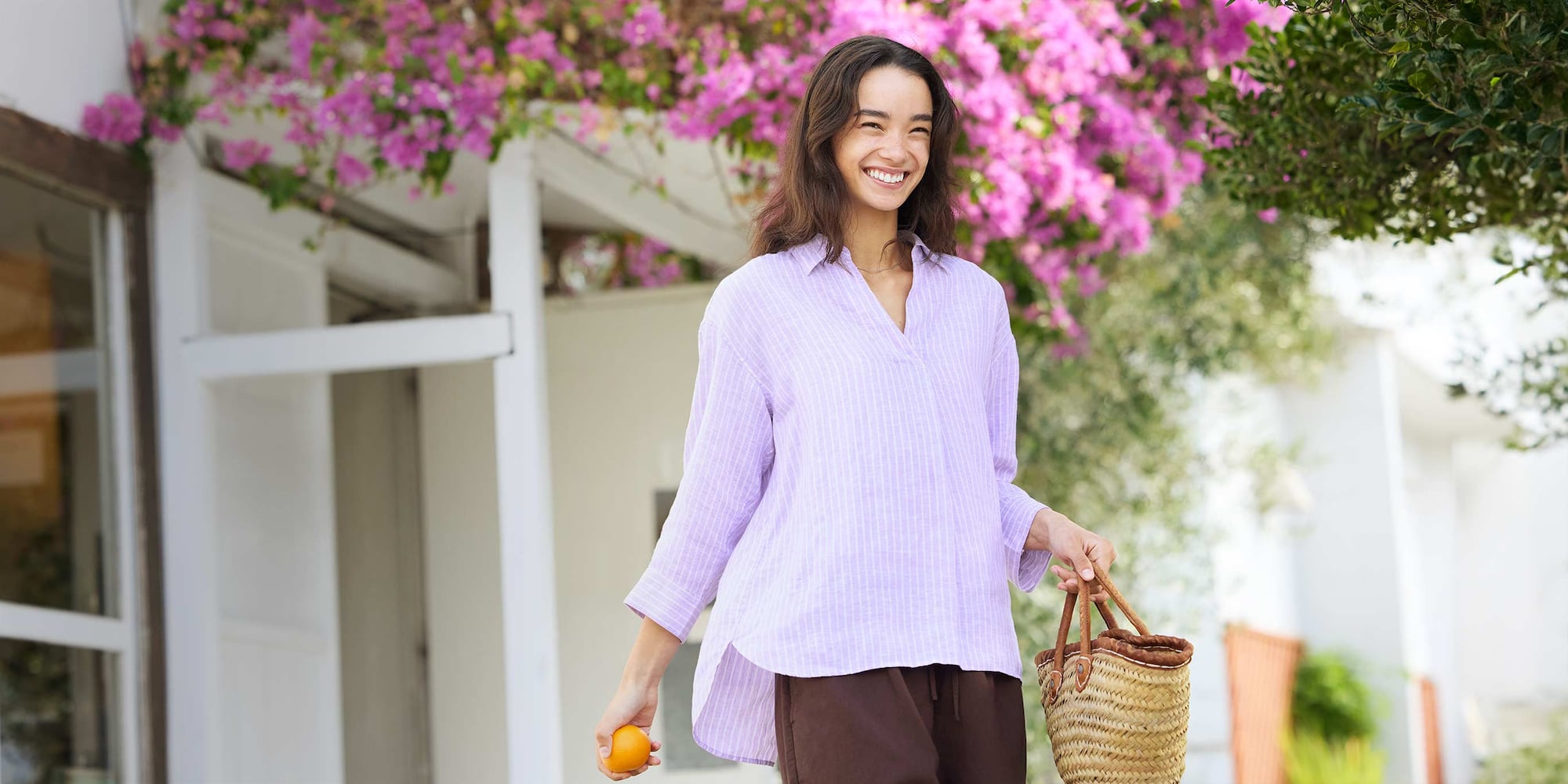model wearing 100% Premium Linen Skipper Collar Shirt in purple stripe with brown trousers while carrying a small basket-style handbag and an orange