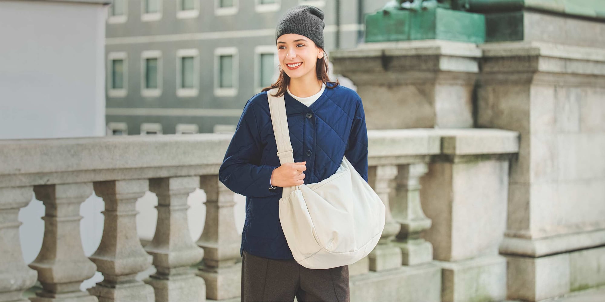 model carrying Puffy Shoulder Bag in natural while wearing a blue quilted jacket over a white crew neck, with brown trousers and a grey beanie
