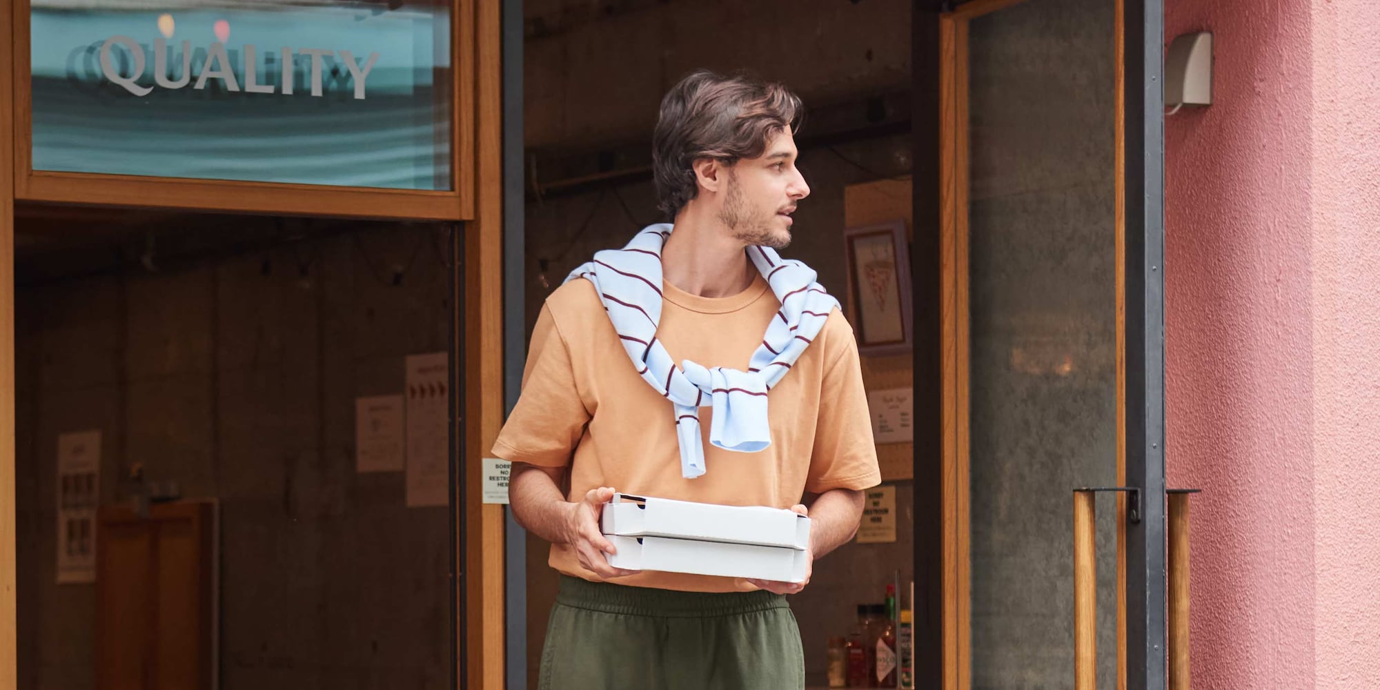 model wearing Crew Neck T-Shirt in orange with khaki trousers and a light blue knit tied around the shoulders while carrying two takeaway pizza boxes