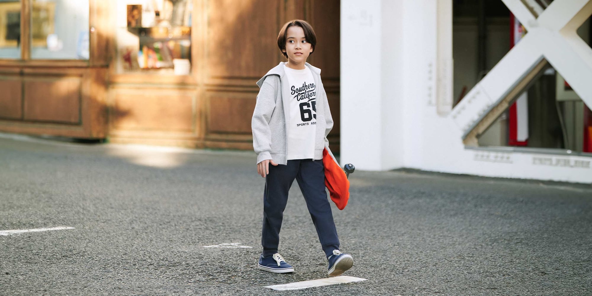 model wearing DRY Sweat Joggers in navy with a white graphic T-shirt and grey hoodie and blue trainers while carrying a skateboard