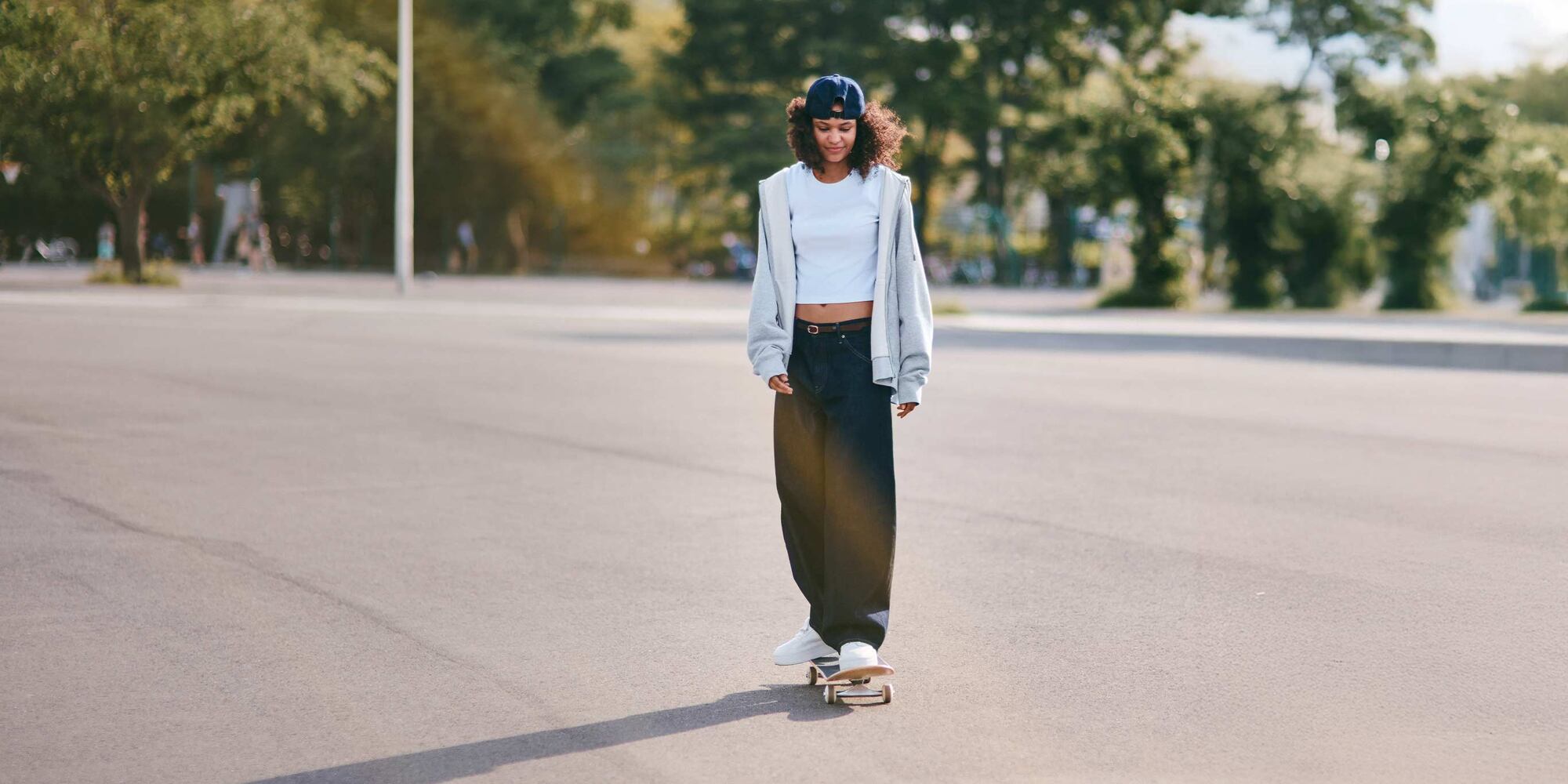 model wearing dark wash Baggy Curve Jeans with a white mini tee, brown belt, light grey hoodie, white trainers and a blue cap while skateboarding