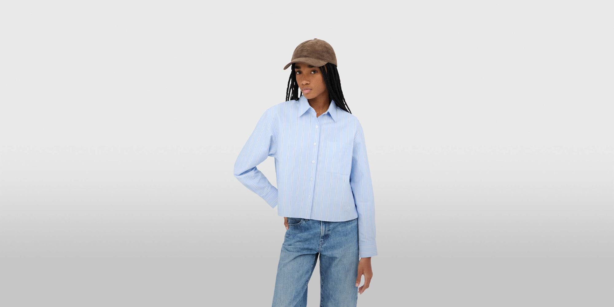 model wearing Oxford Boxy Shirt in blue with mid-blue jeans and a brown cap