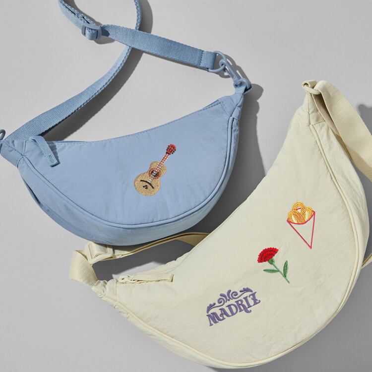 Dos bolsos mini redondeados; uno azul claro con bordado de guitarra, otro color crema con bordado de la palabra Madriz al estilo de los azulejos tradicionales, un clavel rojo y un cucurucho de churros.