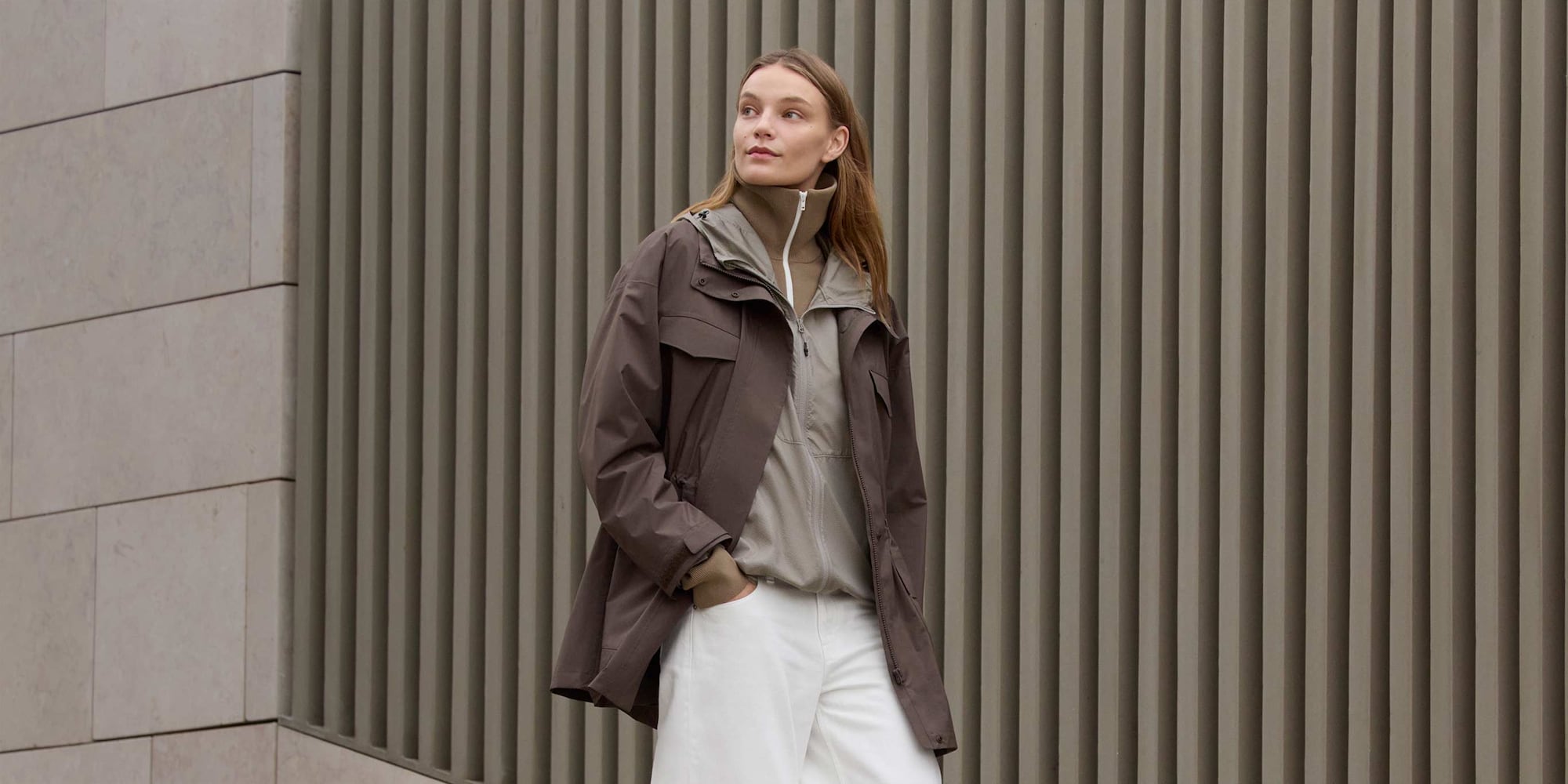 model wearing Utility short coat in brown over a grey jacket and brown zip-up funnel neck top with white trousers