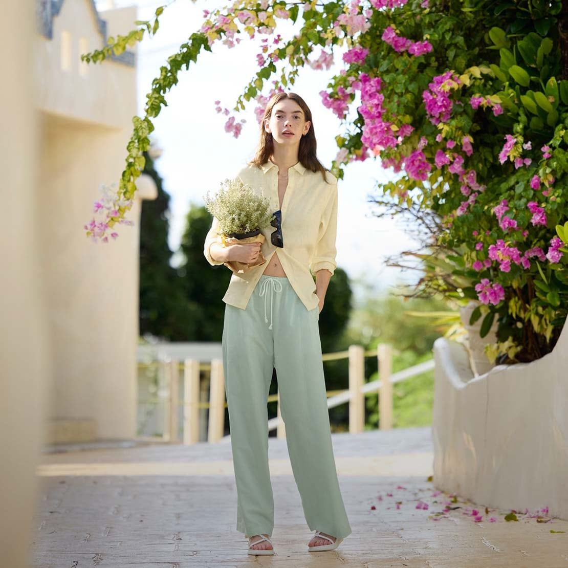 model wearing Linen Blend Easy Trousers in light green with a yellow shirt while holding a potted plant