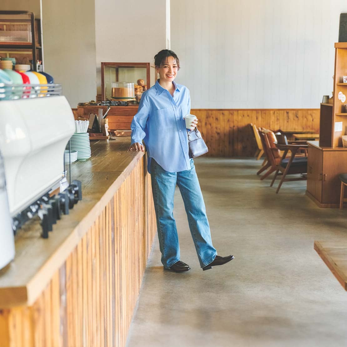 model wearing straight jeans in blue with a light blue shirt and brown loafers standing in a café with a takeaway coffee cup