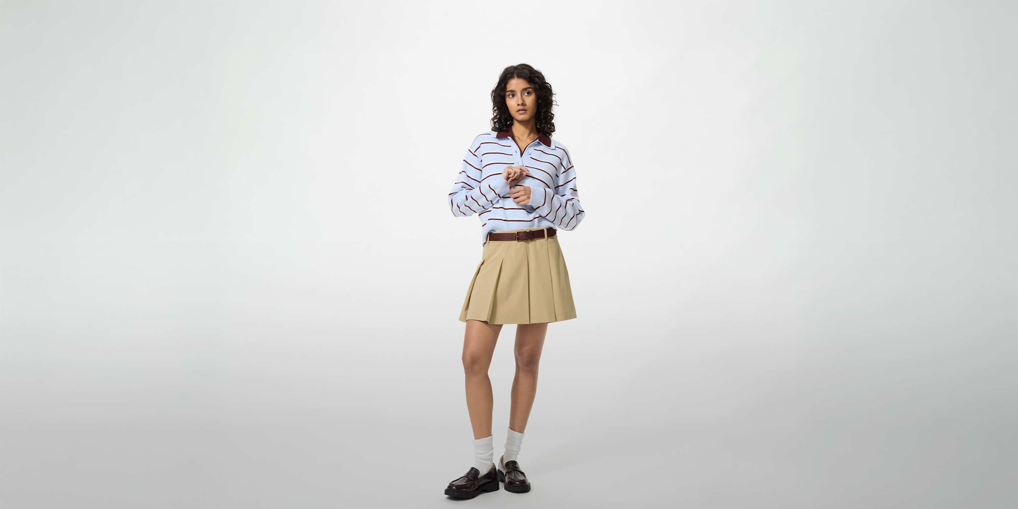 model wearing Pleated Skort in beige with a light blue and maroon knitted polo and a brown belt, white socks and black loafers